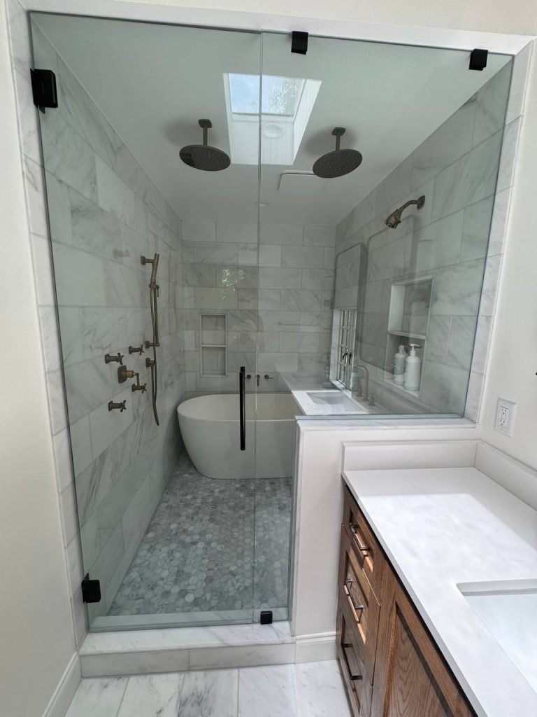Finished glass wet room with two rain shower heads, a freestanding tub, marble-look walls and a pebble mosaic floor.