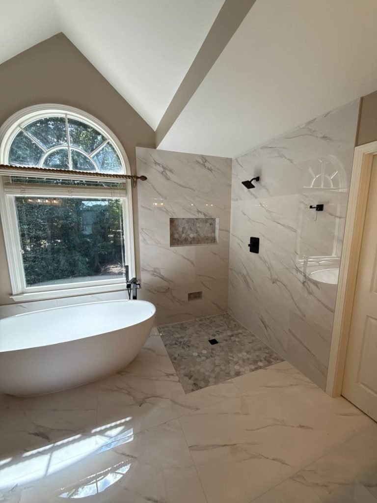 The same master bathroom after restoration: marble-look porcelain wet room with a freestanding tub under an arched window.